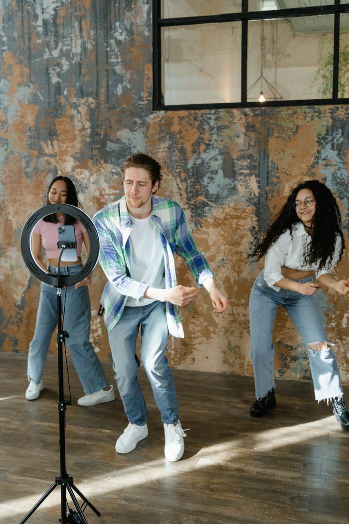 Services Three young adults dancing indoors, creating social media content using a smartphone and ring light.